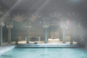 En el interior del Exclusivo MIDRA del Hammam Al Ándalus Málaga se respira un ambiente sereno con aire brumoso, columnas arqueadas y una piscina tranquila iluminada por una suave iluminación ambiental. - ForTwo - Planes para parejas