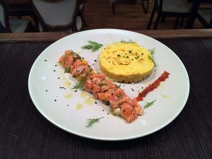 En el Restaurante La Luz de la Candela de Málaga, disfrute de un tartar de salmón servido con una mousse amarilla vibrante y hierbas frescas en un plato blanco impecable. - ForTwo - Planes para parejas