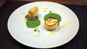 Vieiras selladas con salsa verde y guarnición adornan bellamente un plato blanco, encarnando la esencia de la cocina malagueña en Restaurante La Luz de la Candela - Málaga. - ForTwo - Planes para parejas