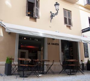 El restaurante La Cocotte en Málaga cuenta con mesas y sillas de madera situadas contra un edificio beige con una elegante lámpara de pared encima, ofreciendo una muestra de cocina internacional. - ForTwo - Planes para parejas