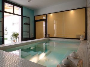 El santuario de Le Patio Spa cuenta con una tranquila piscina de azulejos blancos, rodeada de paredes verdes y faroles. Los amplios ventanales a la izquierda inundan el espacio de luz natural, creando un oasis inspirado en un hammam para una escapada encantadora. - ForTwo - Planes para parejas