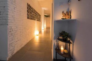 El pasillo, que evoca un rincón sereno de Le Patio Spa, cuenta con una pared de ladrillo blanco iluminada por velas y luces esféricas. Las estanterías exhiben una elegante decoración, que incluye una exuberante planta y delicadas figuras, que invitan a los visitantes a disfrutar de un Ritual Básico con exfoliación en Le Patio Spa. - ForTwo - Planes para parejas