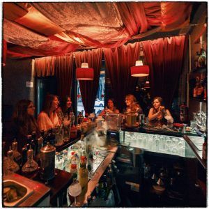 Un acogedor bar en Barcelona, donde la gente se relaja en la barra bajo una luz tenue y cortinas rojas. La selección ofrece una amplia gama de bebidas, incluyendo cócteles exclusivos de su reconocido "Taller de Coctelería: Aprende a Preparar y Degustar 3 Cócteles Exclusivos". - ForTwo - Planes para parejas