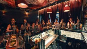 Los visitantes se relajan en una coctelería de Barcelona, disfrutando de la experiencia del Taller de Coctelería donde preparan y saborean 3 cócteles exclusivos bajo una iluminación tenue y cortinas rojas. - ForTwo - Planes para parejas