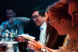 En un animado bar de Barcelona, la gente participa en Taller de Coctelería: Aprende a Preparar y Degustar 3 Cócteles Exclusivos. Botellas y cocteleras se alinean en el mostrador mientras todos sonríen y ríen, disfrutando de esta experiencia única de coctelería. - ForTwo - Planes para parejas