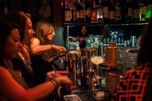 En un acogedor bar barcelonés, con poca luz, la gente disfruta del "Taller de Coctelería: Aprende a Preparar y Degustar 3 Cócteles Exclusivos" mientras alguien sirve bebidas. Botellas y vasos brillan sutilmente al fondo, creando un ambiente acogedor que captura la esencia de la coctelería. - ForTwo - Planes para parejas