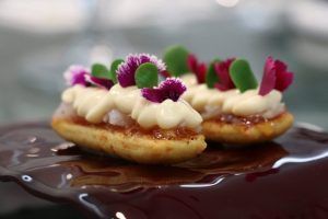 Elegantes eclairs gourmet cubiertos con crema, flores violetas y hojas verdes, que recuerdan el arte de Aire Gastrobar - Cocina mediterránea de autor, reposando sobre una superficie de chocolate brillante. - ForTwo - Planes para parejas
