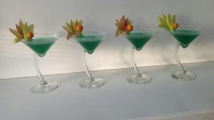 En el Restaurante La Cocotte Cocina Internacional en Málaga, cuatro cócteles verdes en vasos inclinados están elegantemente cubiertos con flores y una esfera naranja, realzando el vibrante estilo internacional. - ForTwo - Planes para parejas