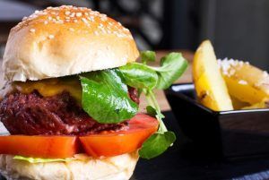 Una jugosa hamburguesa con lechuga, tomate y queso junto con patatas gajo en un plato negro trae el sabor de la cocina internacional del Restaurante La Cocotte directamente a Málaga. - ForTwo - Planes para parejas