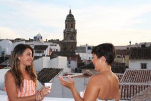 Dos mujeres disfrutan de un momento de tranquilidad en una azotea de Málaga, tomando sus bebidas con el horizonte histórico de la ciudad y la torre del reloj a la vista, evocando la refrescante experiencia del "Baño sencillo en el Hammam Spa de Málaga". - ForTwo - Planes para parejas