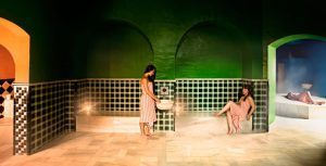 En el ambiente sereno del Hammam Spa, Málaga, las mujeres disfrutan de un momento de tranquilidad mientras una sirve agua a otra sentada con gracia, capturando verdaderamente la esencia de un "Baño sencillo en el Hammam Spa de Málaga". - ForTwo - Planes para parejas