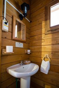 Un baño de estilo rústico con estilo cabaña luce una pared de madera, un elegante lavabo, un espejo y una lámpara. Una pequeña ventana deja entrar la luz natural mientras una toalla cuelga ordenadamente junto al lavabo, evocando momentos de tranquilidad en la Cabaña Águila Imperial, en el Parque Nacional de Cabañeros. - ForTwo - Planes para parejas