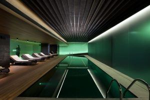 Relájese en la tranquila piscina cubierta del Mandarin Hotel en Barcelona SPA, que cuenta con tumbonas en una terraza de madera, iluminación moderna y paneles de techo oscuros en ángulo que evocan la elegancia arquitectónica de Barcelona: una escapada perfecta tipo spa. - ForTwo - Planes para parejas