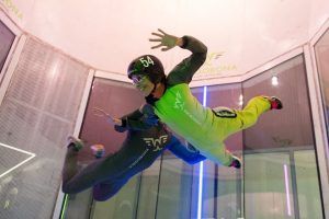 Dos personas experimentan "Paracaidismo en túnel de viento Windobona Indoor Skydiving en Madrid", volando en un túnel de viento vertical con cascos y trajes de vuelo, con un fondo brillante como telón de fondo. - ForTwo - Planes para parejas