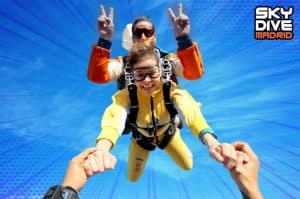 Dos personas sonríen y se toman de la mano en el aire, disfrutando de la emoción de un Salto en Paracaídas con Skydive Madrid contra un cielo azul intenso, con el logotipo de Skydive Madrid brillando detrás de ellos. - ForTwo - Planes para parejas