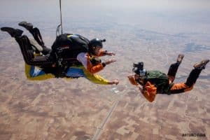 Tres paracaidistas caen libremente sobre un vasto paisaje, y uno de ellos captura la impresionante escena con una cámara, resaltando la emoción de un "Salto en paracaídas con Skydive Madrid". - ForTwo - Planes para parejas