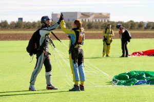 Dos paracaidistas celebran chocando los cinco en un campo de hierba en Skydive Madrid, con sus paracaídas y equipo a su lado, mientras otros en el fondo recogen sus cosas después de su emocionante "Salto en paracaídas con Skydive Madrid". - ForTwo - Planes para parejas