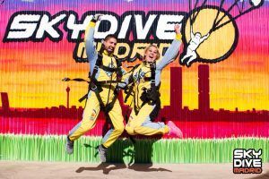 Dos personas con equipo de paracaidismo saltan alegremente frente a un vibrante telón de fondo de Skydive Madrid, celebrando su emocionante experiencia con "Salto en paracaídas con Skydive Madrid". - ForTwo - Planes para parejas