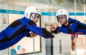 Dos personas con trajes de vuelo azules y cascos practican paracaidismo indoor en túnel de viento Madrid Fly, sonriendo ampliamente mientras se miran uno al otro. - ForTwo - Planes para parejas