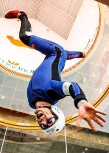 Vestido con traje azul y casco, alguien salta alegremente boca abajo en Madrid Fly, experimentando el paracaidismo indoor en el túnel de viento. - ForTwo - Planes para parejas