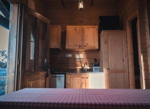 La acogedora cocina de una cabaña de madera cuenta con la luz del sol que entra a través de una puerta abierta y una mesa cubierta con un mantel a cuadros rojos, ofreciendo la calidez y el encanto de "Cabaña en el árbol para ir en pareja en MonteHoliday Madrid", perfecta para parejas que buscan tranquilidad. - ForTwo - Planes para parejas