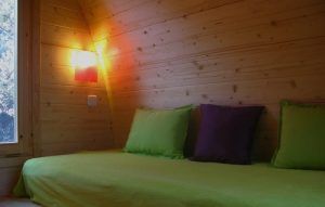 Relájese en una acogedora habitación de madera en la Cabaña en el árbol para ir en pareja en MonteHoliday Madrid, que cuenta con una colcha verde, almohadas verdes y moradas y una cálida iluminación de pared junto a la ventana. - ForTwo - Planes para parejas