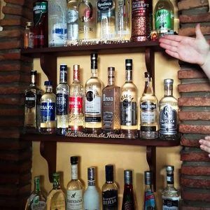 Los estantes del Restaurante La Alacena de Francis en Málaga exhiben una variedad de botellas de vodka sobre una rústica pared de ladrillo. Dos manos invitan a la selección, capturando la esencia de este encantador local. - ForTwo - Planes para parejas