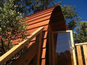 Escápate a la Cabaña en el árbol para ir en pareja en MonteHoliday Madrid, una encantadora cabaña de madera con techo curvo, situada entre árboles bajo un cielo azul claro, perfecta para parejas que buscan tranquilidad en la naturaleza. - ForTwo - Planes para parejas