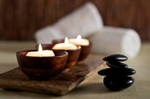 Velas encendidas en soportes de madera sobre una bandeja, con piedras negras apiladas y toallas blancas enrolladas, evocan un ambiente de spa que recuerda a la Vinoterapia en el spa urbano Ambrosia en Barcelona, ideal para disfrutar de una Vinoterapia relajante. - ForTwo - Planes para parejas
