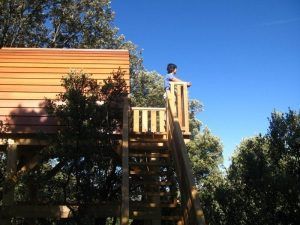 Una pareja disfruta de una terraza de madera en la Cabaña en el árbol para ir en pareja en MonteHoliday Madrid, rodeada de exuberante vegetación y un cielo azul claro. - ForTwo - Planes para parejas