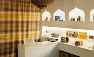 El Hammam Ritual de los Sultanes en Rituels d'Orient Barcelona cuenta con una acogedora recepción de spa con estanterías en forma de arco, productos para el cuidado de la piel y una cortina de rayas. La cálida iluminación realza el ambiente, capturando la esencia de Rituels d'Orient. - ForTwo - Planes para parejas