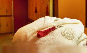 Vestido con una bata blanca con el logo bordado y una borla roja, te deleitas con el cálido resplandor del Hammam Ritual de los Sultanes en Rituels d'Orient Barcelona, entre susurrantes taquillas de madera. - ForTwo - Planes para parejas