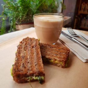 Experimenta el ambiente perfecto de un brunch en MAAI Barcelona: un sándwich tostado con relleno derretido sobre papel de horno, acompañado de café espumoso, tenedor y cuchillo. Disfruta del ambiente de comida local, enmarcado por una planta de fondo. - ForTwo - Planes para parejas
