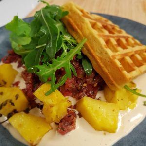 Disfrute de un delicioso brunch en MAAI Barcelona: un gofre con tartar de ternera, rúcula y trozos de piña, bañado en salsa cremosa, servido en un plato oscuro. Producto: Brunch de comida local en MAAI Barcelona. - ForTwo - Planes para parejas