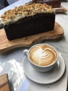 Un capuchino con arte latte reposa junto a un bizcocho de chocolate cubierto de nueces en la mesa de mármol, ideal para un relajado gastro-brunch en Enkel Barcelona. - ForTwo - Planes para parejas