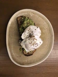 El gastro-brunch en Enkel Barcelona incluye una tostada de aguacate con dos huevos poché y semillas, elegantemente servida en un plato de cerámica. Colocada sobre una mesa de madera, ofrece sabor y estilo. - ForTwo - Planes para parejas