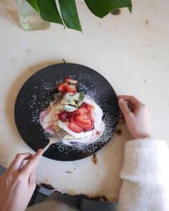 Una persona disfruta de un brunch de panqueques cubiertos con fresas, crema y menta en el restaurante Brunch en Picnic de Barcelona, servido en un plato negro en una mesa blanca. - ForTwo - Planes para parejas