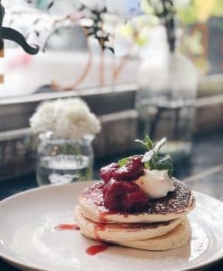 En el restaurante Brunch en Picnic de Barcelona, panqueques aderezados con fresas, crema batida y menta se presentan elegantemente en un plato blanco en medio de un acogedor ambiente de cafetería. - ForTwo - Planes para parejas