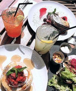 El brunch en el restaurante Brunch en Picnic de Barcelona incluye panqueques decorados artísticamente, tostadas francesas, ensalada y bebidas frutales en una mesa de madera iluminada por el sol. - ForTwo - Planes para parejas