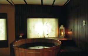Una acogedora sala de spa con bañera de hidromasaje de madera, linternas iluminadas con velas e iluminación suave en una pared texturizada crea el ambiente perfecto para el relajante "Spa y masaje a lo japonés + menú de tapas en Madrid". - ForTwo - Planes para parejas