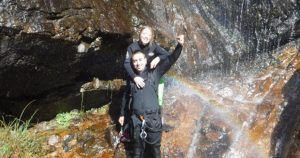 Una pareja con traje de neopreno posa alegremente bajo una cascada en Somosierra, Madrid, con un arcoíris sobre sus cabezas. Uno lleva al otro juguetonamente a cuestas, encarnando el espíritu aventurero de su experiencia de barranquismo en pareja. - ForTwo - Planes para parejas