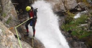 Una persona con casco y traje de neopreno disfruta del barranquismo con el paquete "Barranquismo en pareja en Somosierra - Madrid", descendiendo con destreza una cascada con una cuerda verde en el cautivador paisaje de Somosierra. - ForTwo - Planes para parejas