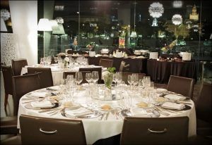 En el suavemente iluminado Restaurante Abades Triana, las mesas elegantemente dispuestas con platos, vasos y cubiertos ofrecen una vista impresionante del paisaje urbano por la noche. - ForTwo - Planes para parejas