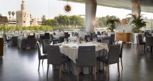 El Restaurante Abades Triana presume de un elegante interior con mesas redondas y sillas grises. Sus amplios ventanales ofrecen impresionantes vistas al río, a una torre y a los frondosos árboles del encantador barrio de Triana. - ForTwo - Planes para parejas