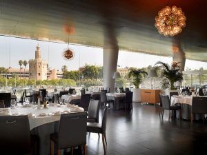 El Restaurante Abades Triana combina el encanto moderno con grandes ventanales que ofrecen vistas a una torre histórica y una exuberante vegetación, proporcionando mesas elegantemente dispuestas para una experiencia gastronómica inolvidable. - ForTwo - Planes para parejas