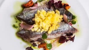 En el Restaurante La Rotonda del Hotel Westin Palace Madrid, saboree un plato de pescado cubierto con frutas amarillas cortadas en cubitos, adornado con verduras frescas y vegetales coloridos en un plato blanco impecable. - ForTwo - Planes para parejas