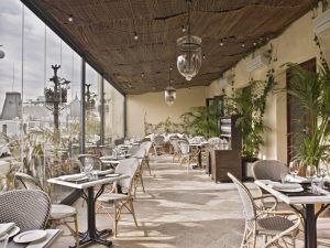 El Restaurante Ático del Hotel The Principal Madrid cuenta con una elegante terraza con sillas de mimbre, mesas de cristal y lámparas colgantes, todo ello ofreciendo una encantadora vista de la ciudad en el corazón de Madrid. - ForTwo - Planes para parejas