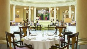 Un lujoso comedor en el Restaurante La Rotonda del Hotel Westin Palace Madrid, con un mantel blanco, rodeado de sillas en una espaciosa sala con columnas y centros de mesa florales. - ForTwo - Planes para parejas