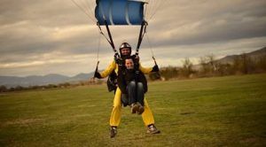 Dos personas hacen paracaidismo en tándem y aterrizan suavemente en un campo de hierba bajo las nubes. Una lleva un traje amarillo brillante. ¡Vive la emoción del salto en tándem de paracaidismo con Skydive Empuriabrava!. - ForTwo - Planes para parejas