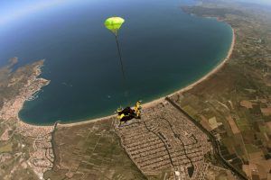 Un paracaidista con un paracaídas verde se desliza sobre la ciudad costera y el océano en un día claro, emocionándose en un Salto tándem de paracaidismo con Skydive Empuriabrava, mostrando impresionantes paisajes debajo. - ForTwo - Planes para parejas